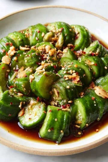 A plate of Asian-style cucumber salad with sliced cucumbers coated in a dark soy-based dressing, garnished with crushed peanuts and red pepper flakes.