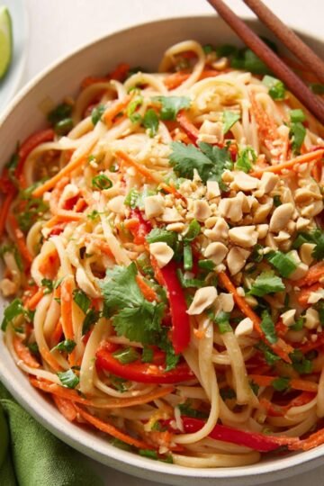 Bowl of Asian noodle salad with rice noodles, shredded carrots, red bell peppers, chopped peanuts, cilantro, and lime wedges on the side, served with chopsticks.