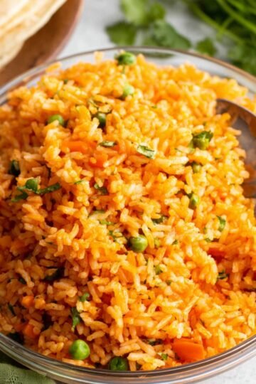 A clear glass bowl of cooked Authentic Mexican Rice with peas and carrots, garnished with chopped cilantro, with a serving spoon resting inside.