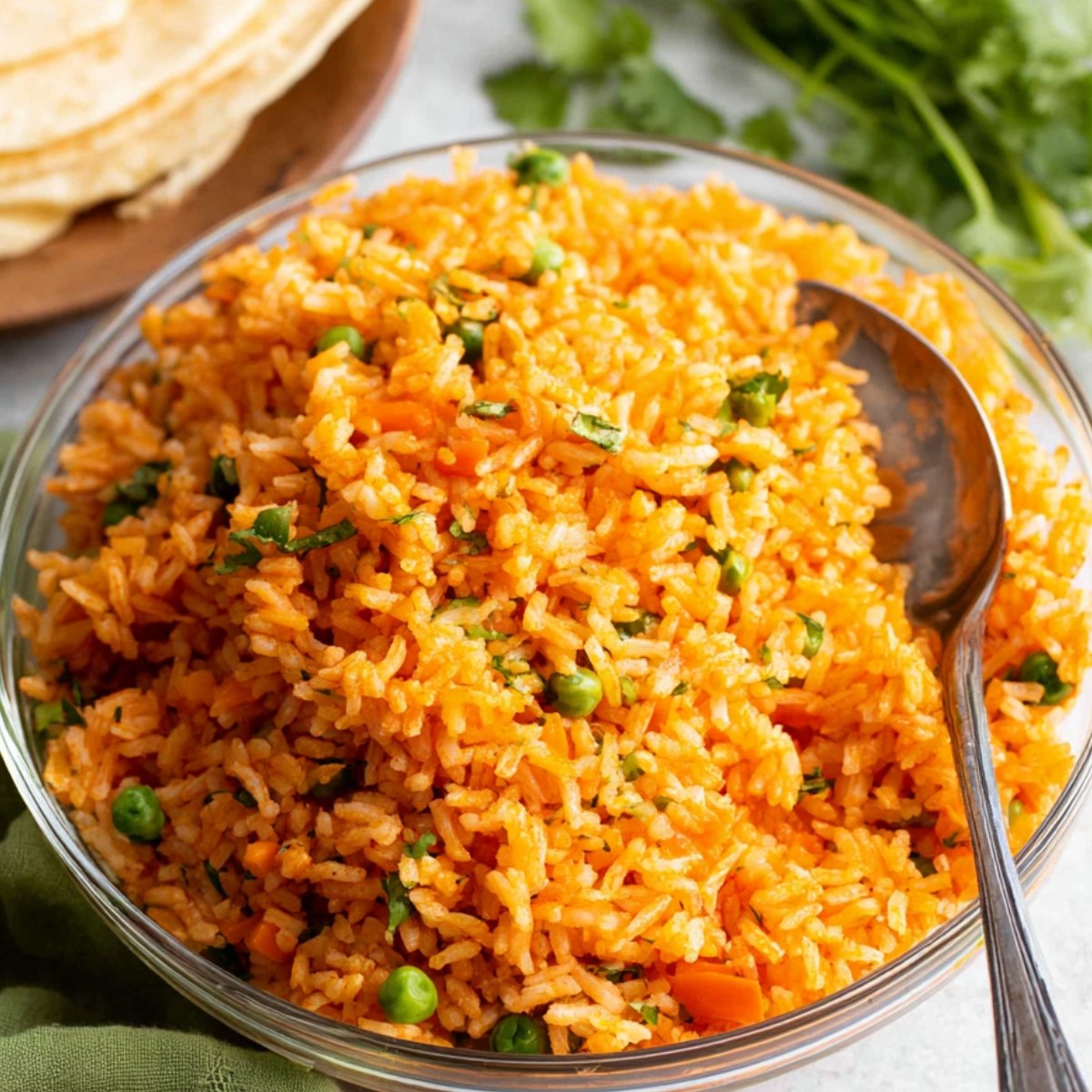 A clear glass bowl of cooked Authentic Mexican Rice with peas and carrots, garnished with chopped cilantro, with a serving spoon resting inside.
