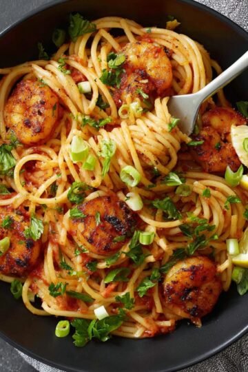 A black bowl filled with spaghetti pasta tossed with blackened shrimp in a tomato-based sauce, garnished with chopped parsley and sliced green onions, with two lemon wedges on the side and a fork resting in the pasta, overhead view.