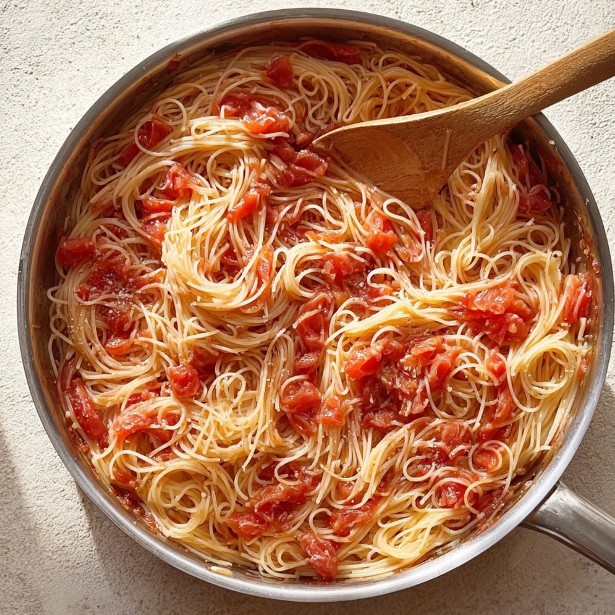 Overhead view of a stainless steel skillet filled with thin spaghetti noodles mixed with a simple tomato sauce, a wooden spoon resting in the pan, bright natural lighting highlighting the glossy texture of the pasta and chunks of tomato.