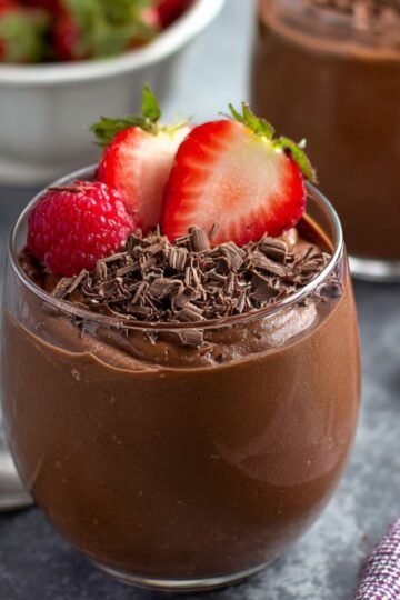 A glass filled with creamy Blended Chocolate Chia Pudding, topped with fresh strawberry halves, a raspberry, and chocolate shavings, placed on a dark surface with a blurred background of more berries and pudding.
