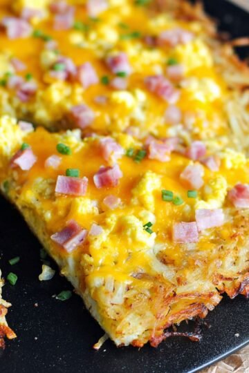 A sliced Breakfast Pizza with Hash Brown Crust on a black pan, showing the golden, crispy hash brown crust, melted cheddar cheese, scrambled eggs, diced ham, and garnished with chopped green onions.