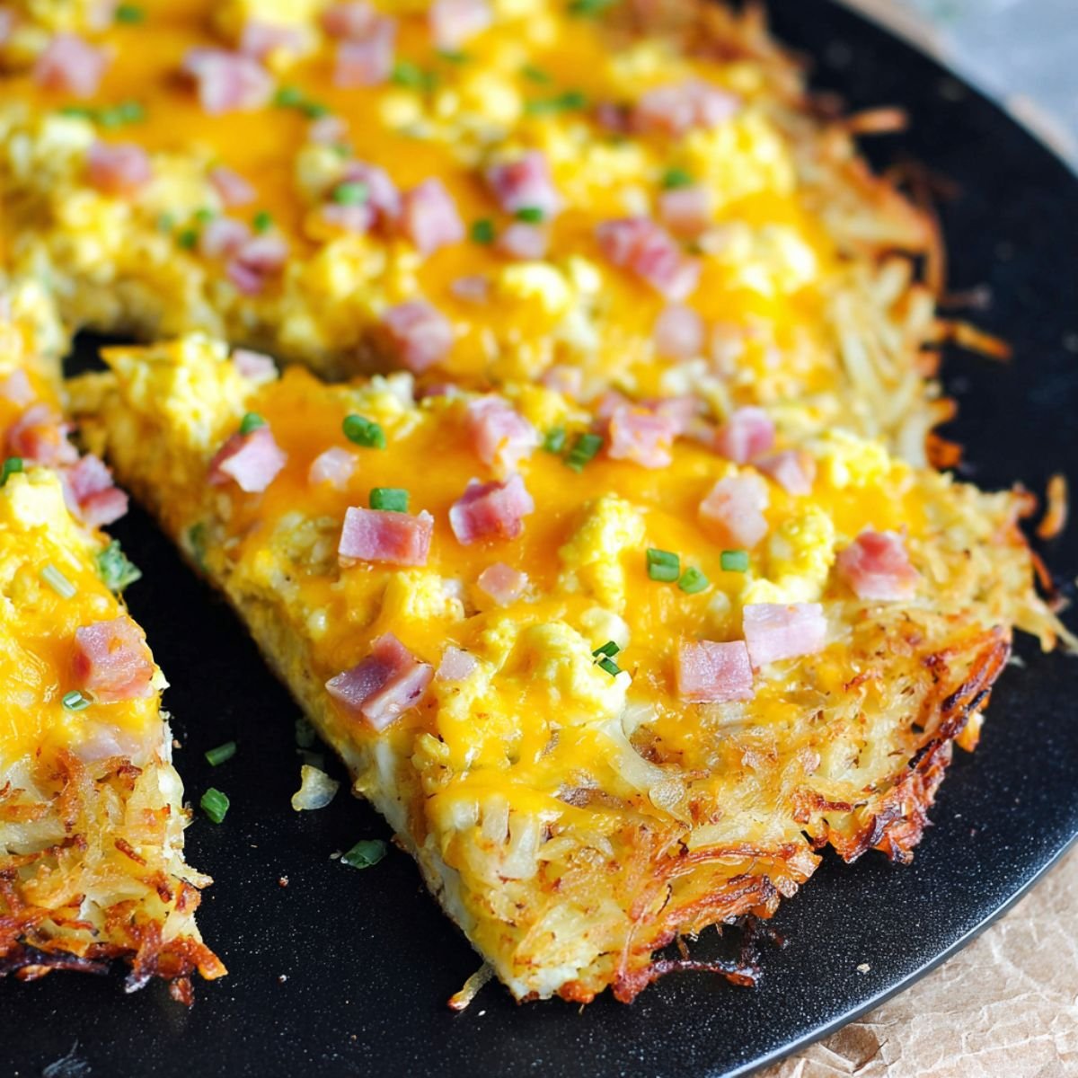 A sliced Breakfast Pizza with Hash Brown Crust on a black pan, showing the golden, crispy hash brown crust, melted cheddar cheese, scrambled eggs, diced ham, and garnished with chopped green onions.