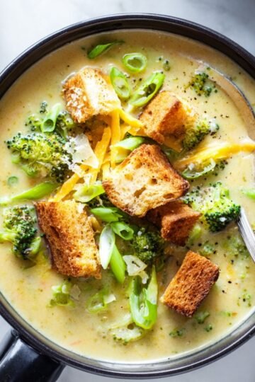 A bowl of creamy soup with chunks of broccoli, shredded cheddar cheese, croutons, and sliced green onions, with a spoon in the bowl.
