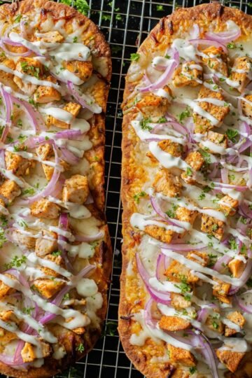 Two baked Buffalo Chicken French Bread Pizza topped with buffalo chicken, melted cheese, sliced red onions, and drizzled with ranch dressing, garnished with chopped parsley.