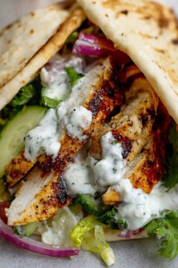 A close-up of a chicken shawarma wrap with sliced grilled chicken, fresh lettuce, cucumber, tomato, red onion, and a drizzle of creamy white sauce, wrapped in a flatbread.