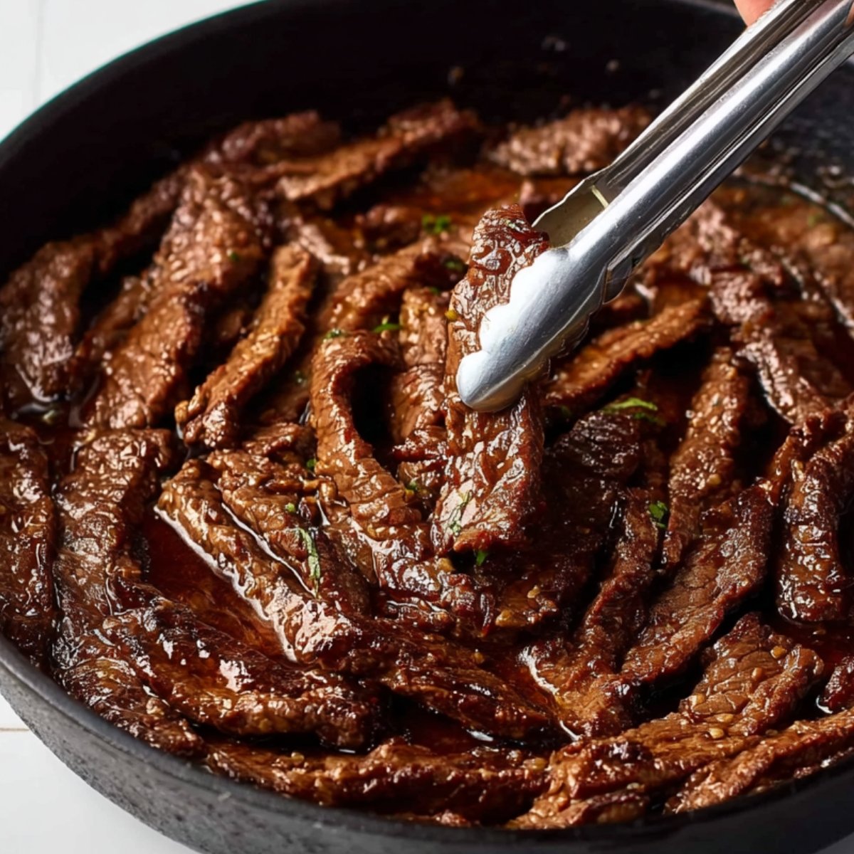 A close-up of a skillet with cooked, sliced beef strips being held with tongs, glistening in a savory sauce.