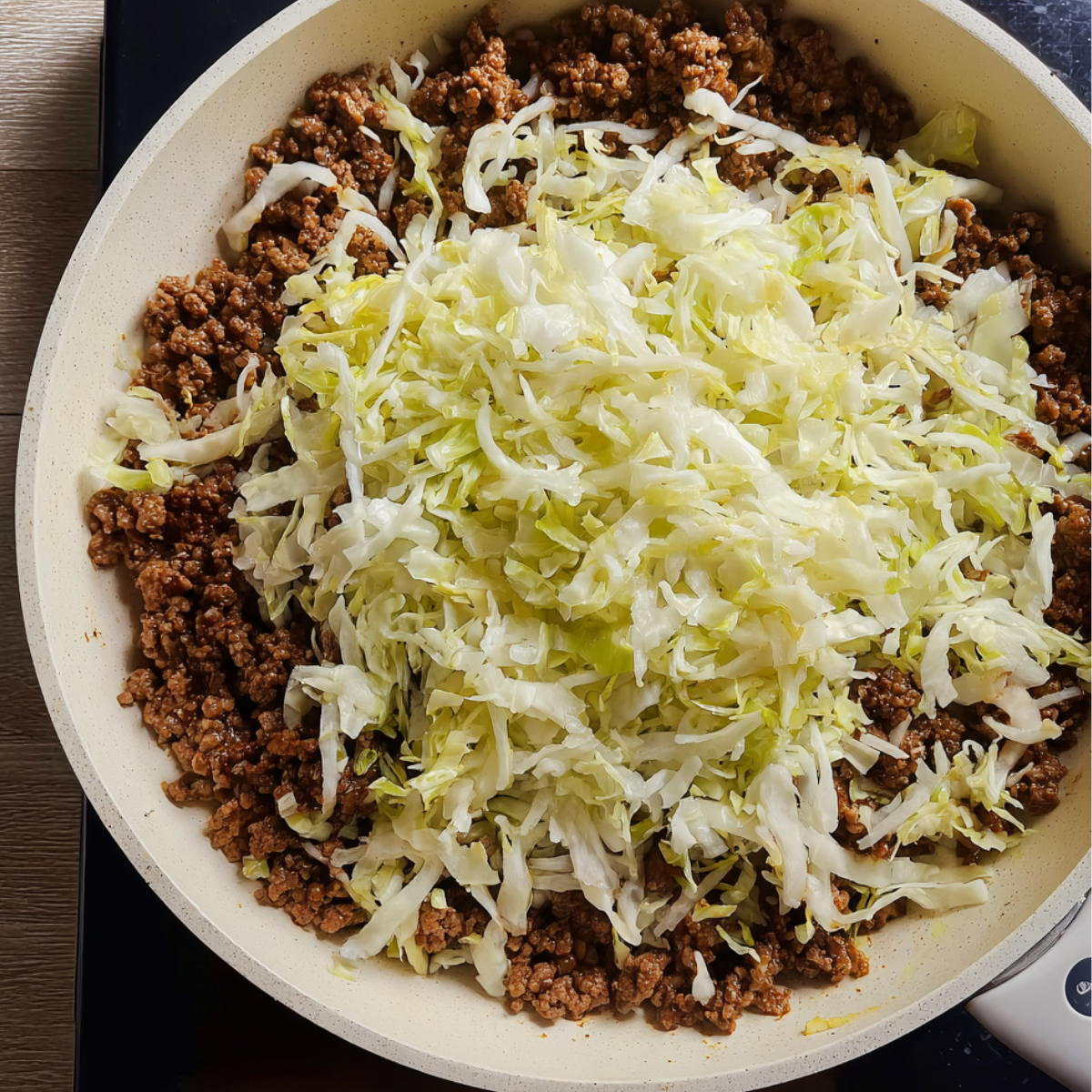 A top-down view of a pan filled with cooked ground beef topped with a generous layer of shredded cabbage, not yet mixed in.