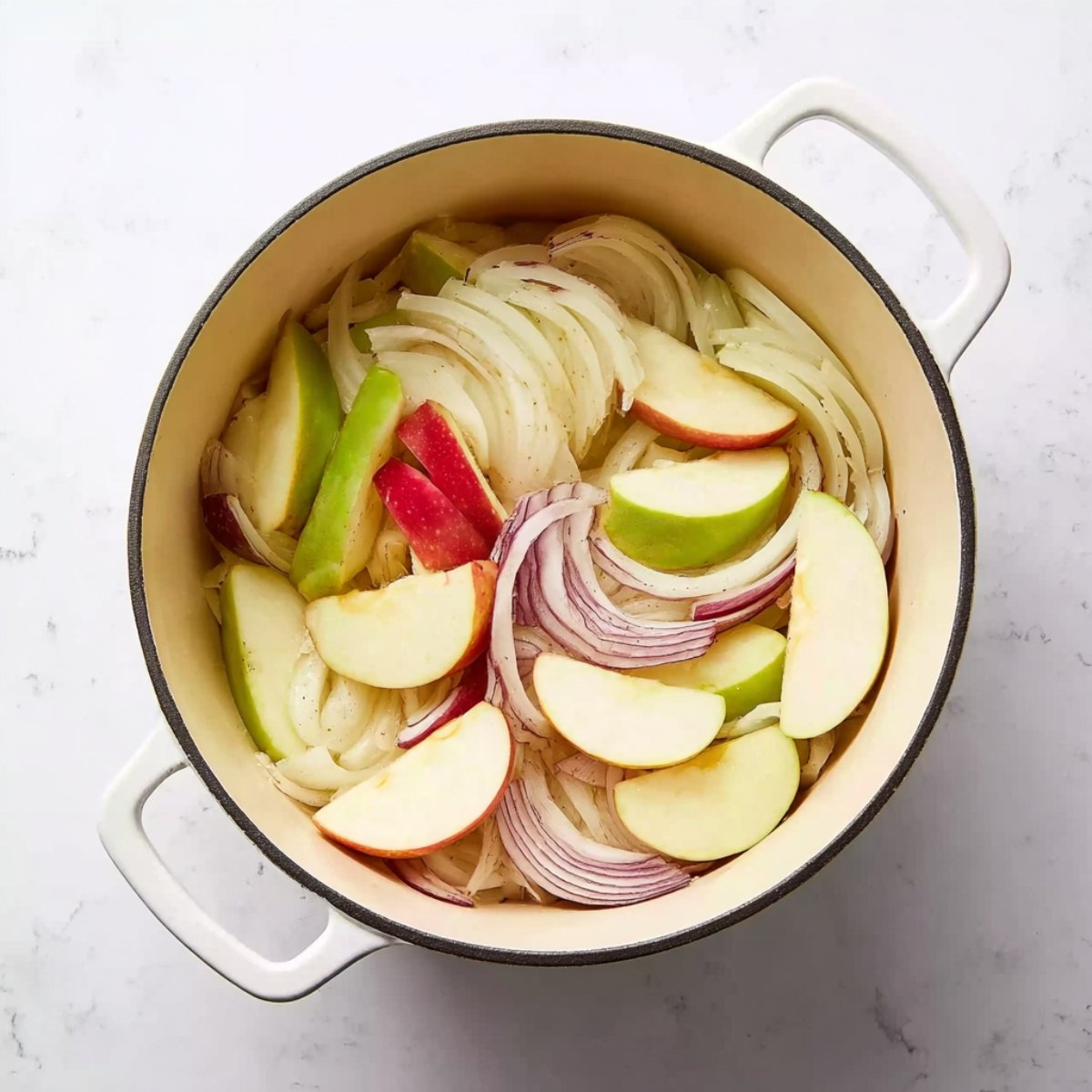Overhead view of a white Dutch oven containing sliced onions and wedges of red and green apples, arranged evenly and ready for cooking.