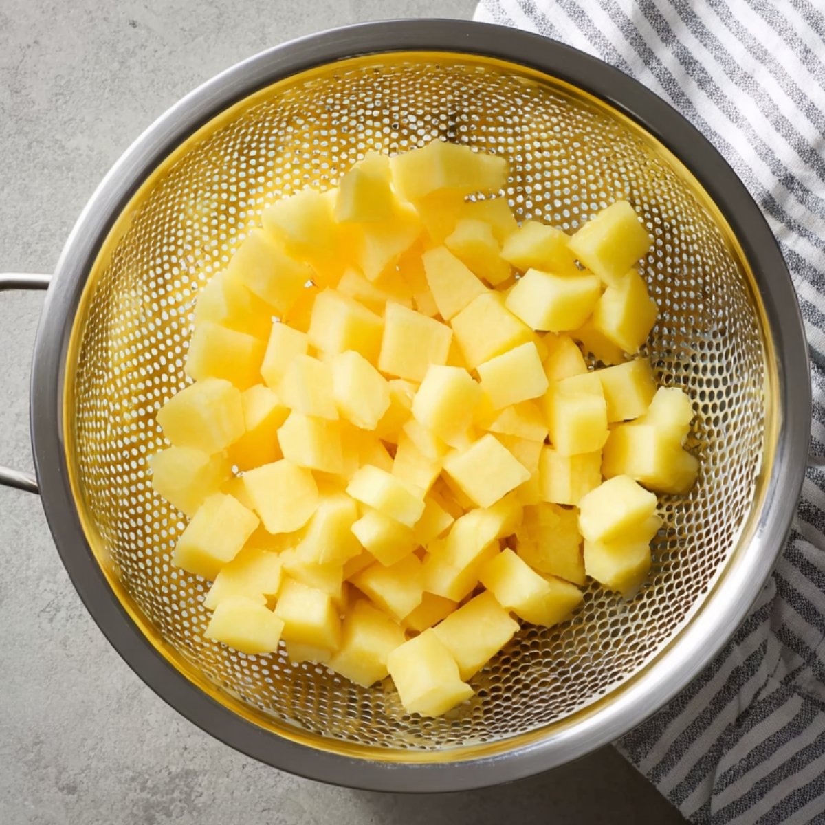 Overhead view of a metal colander containing diced cooked yellow potatoes, placed on a gray countertop with a striped kitchen towel.