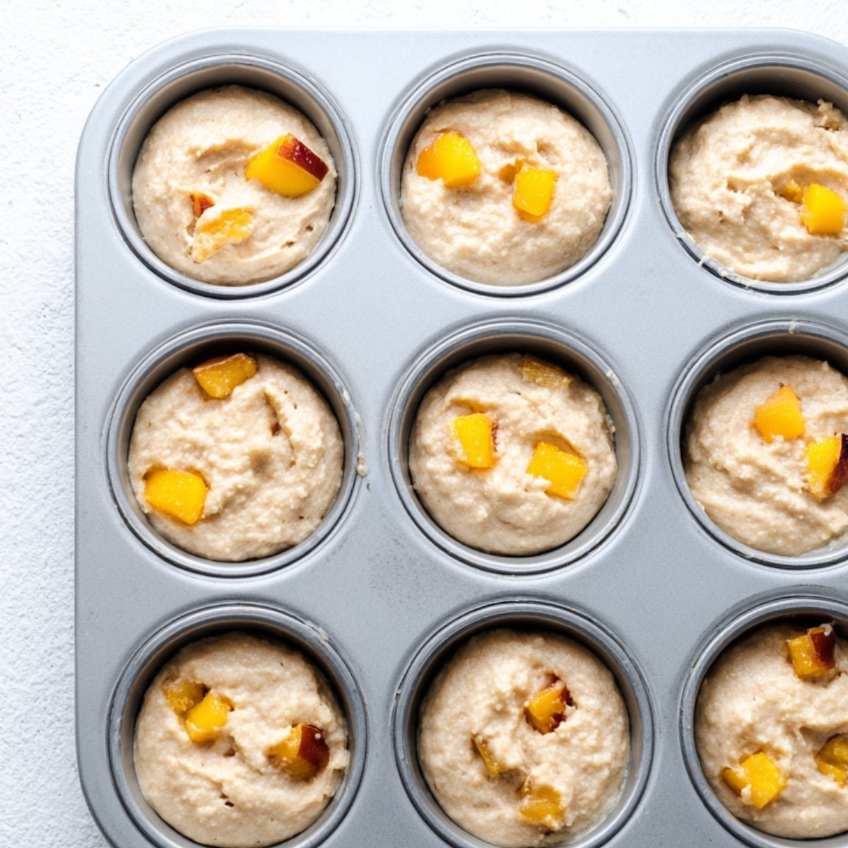 Unbaked muffin batter in a 12-cup muffin tin, each cup containing small chunks of peaches mixed into the batter
