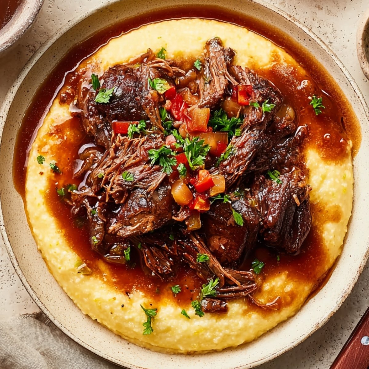 A bowl of Italian-style pot roast served over creamy polenta. The shredded beef is topped with a rich brown sauce and diced vegetables, garnished with fresh parsley.
