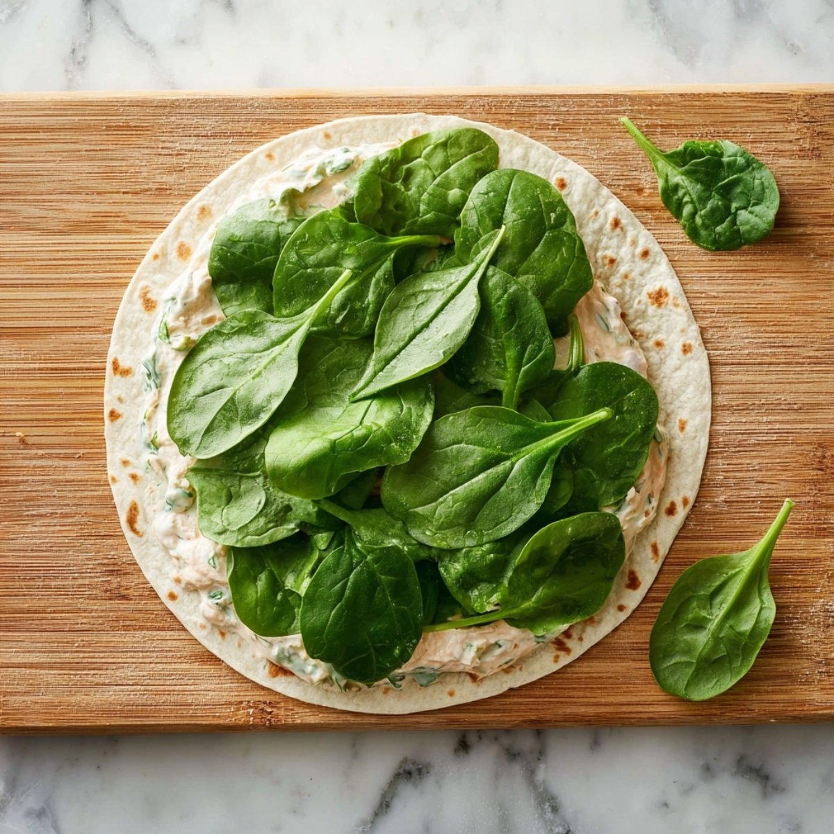 Flour tortilla spread with creamy filling and topped with fresh spinach leaves on a wooden cutting board.