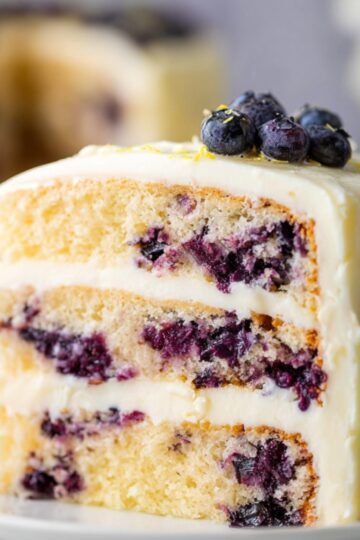 Slice of layered lemon blueberry cake on a plate, with frosting between layers and fresh blueberries on top