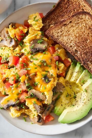 A plate of Loaded Scrambled Eggs mixed with mushrooms, diced tomatoes, and melted cheddar cheese, served with sliced avocado and two pieces of toasted whole-grain bread, garnished with chopped chives.