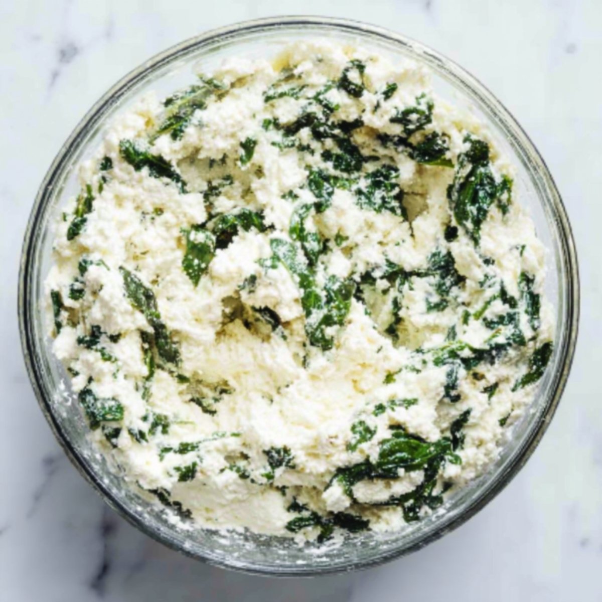 A glass bowl filled with ricotta cheese mixed with chopped spinach, ready to be used as a filling for pasta