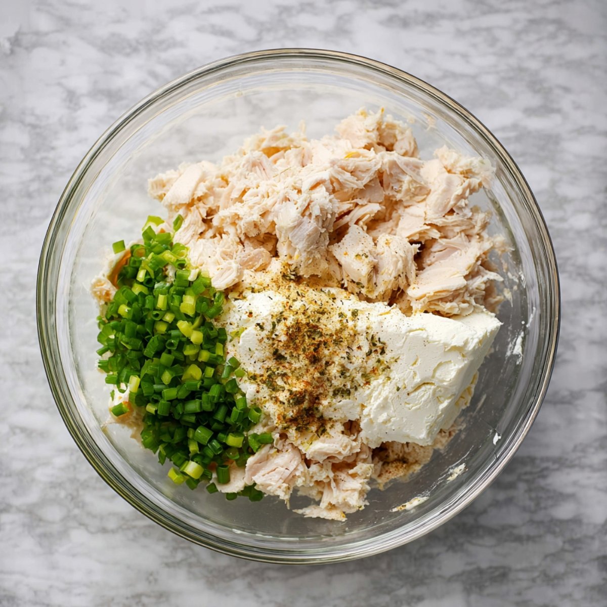 Glass bowl containing shredded chicken, cream cheese, chopped green onions, and seasoning, ready to be mixed