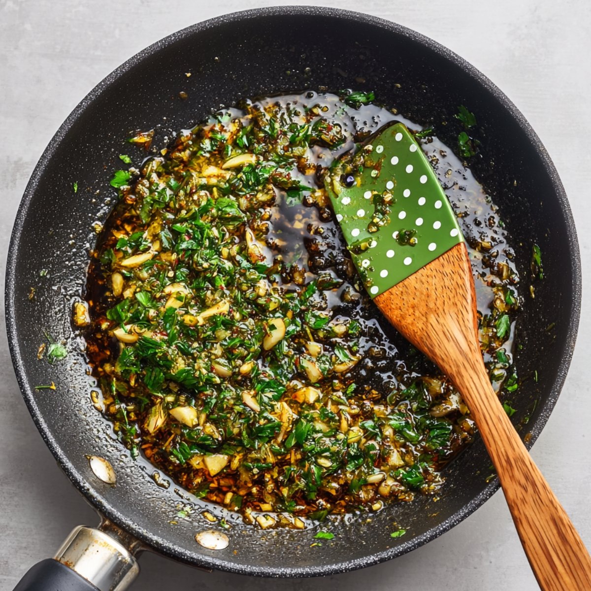 Overhead view of a black skillet with herb oil being cooked, containing chopped garlic and fresh herbs, with a green spatula resting in the pan.