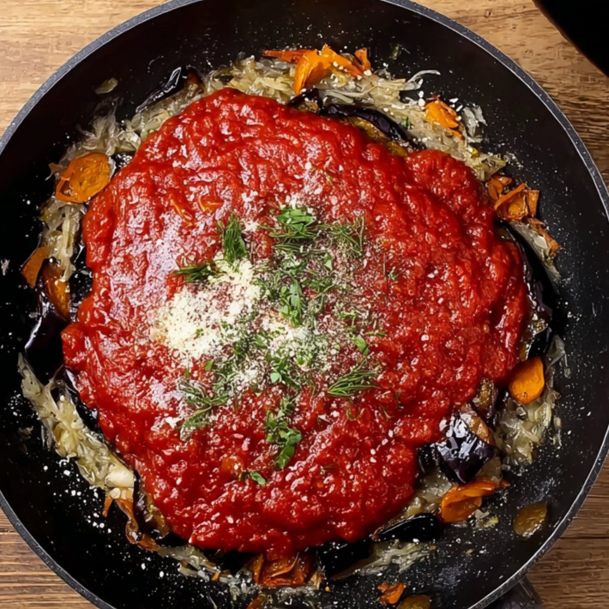 A black skillet with sautéed vegetables topped with bright red tomato sauce, sprinkled with herbs and grated cheese