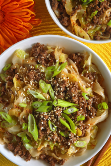 Two white bowls filled with cooked Mongolian Ground Beef and Cabbage, garnished with sliced green onions and sesame seeds, placed on a bright yellow cloth with an orange flower beside them.