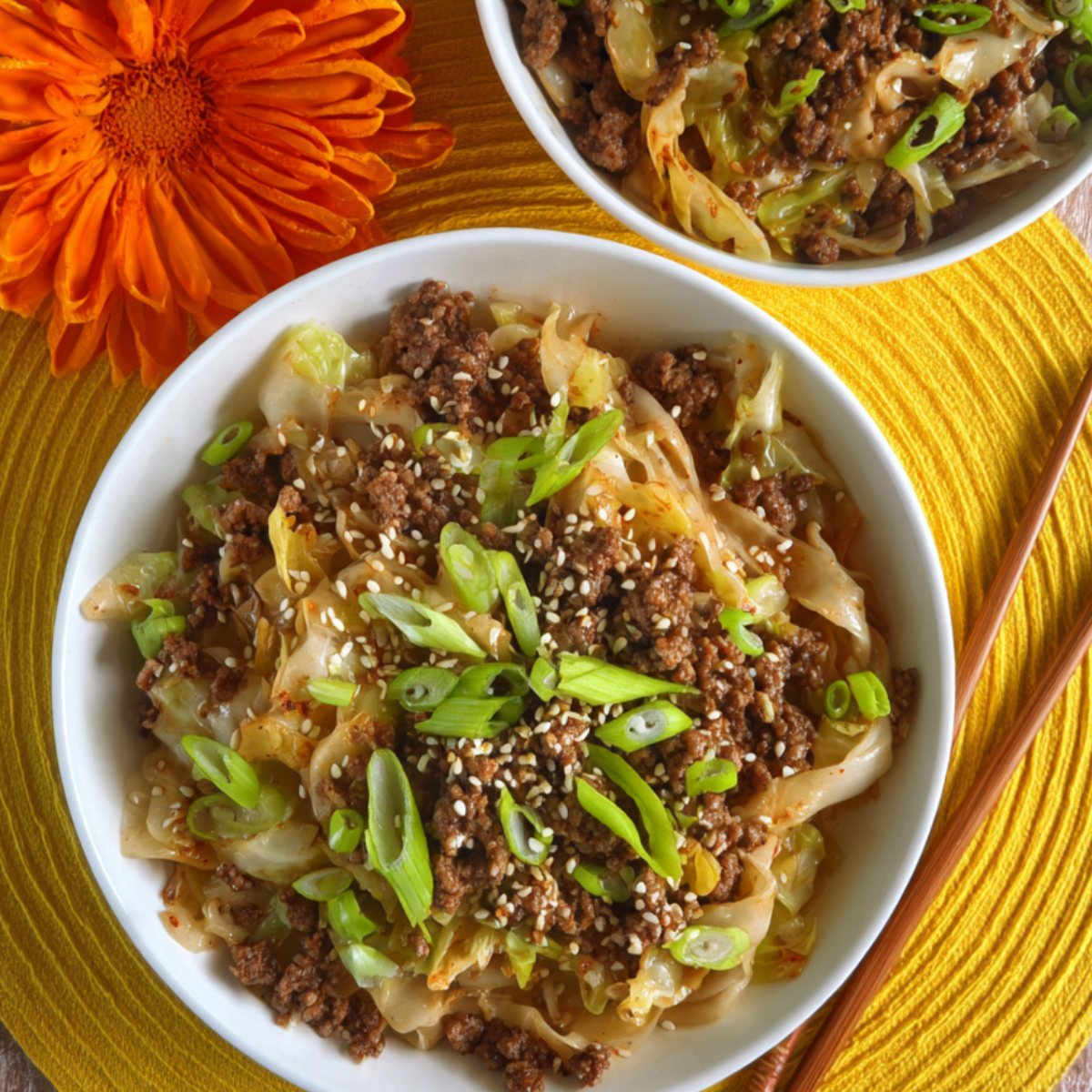 Two white bowls filled with cooked Mongolian Ground Beef and Cabbage, garnished with sliced green onions and sesame seeds, placed on a bright yellow cloth with an orange flower beside them.