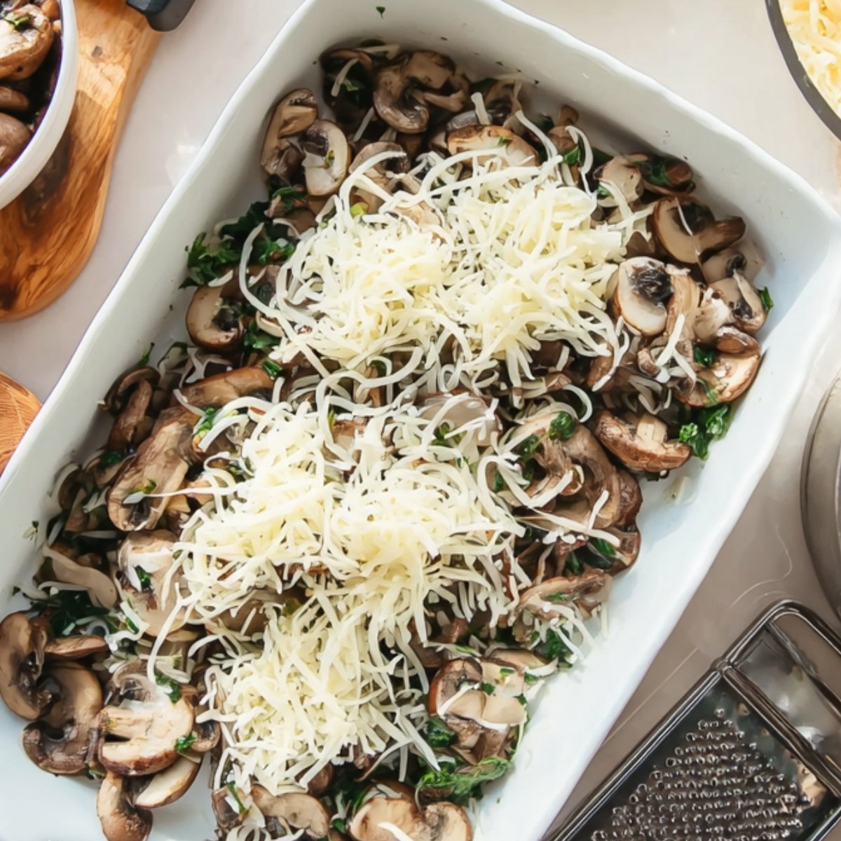 A white rectangular baking dish filled with layers of spinach, mushrooms, and shredded cheese, prepared and ready to bake.
