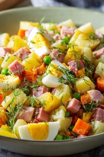 Close-up of a bowl of Olivier Potato Salad with diced potatoes, carrots, peas, ham, and hard-boiled eggs, garnished with fresh dill.