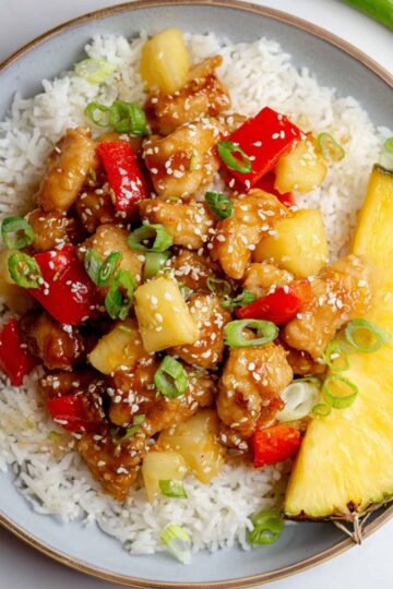 Pineapple Chicken and Rice stir-fry served over white rice on a plate, garnished with green onions and sesame seeds, with a pineapple slice on the side