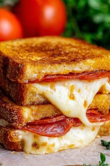 Close-up of a stacked Pizza Grilled Cheese sandwich with melted mozzarella and slices of pepperoni, showing golden toasted bread and gooey cheese stretching slightly.
