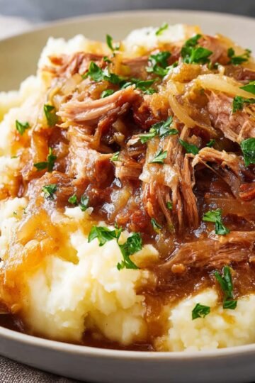 Close-up of shredded, cooked pork served over mashed potatoes, topped with caramelized onions and garnished with chopped parsley.