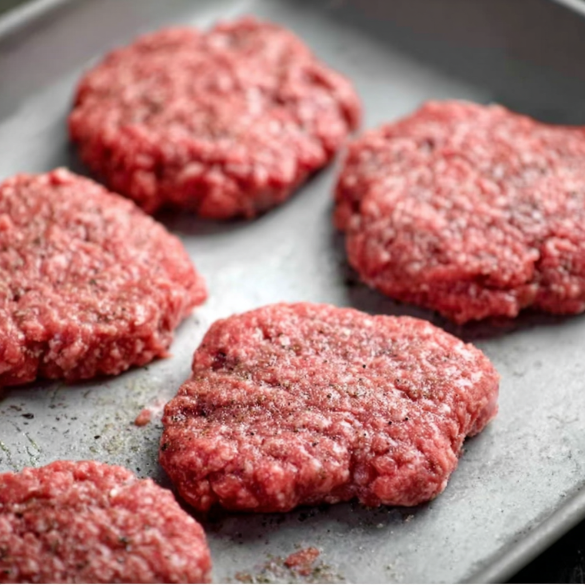 Six raw hamburger patties made from ground beef arranged on a metal baking sheet, ready to be cooked.