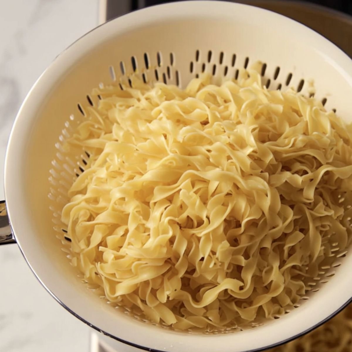 A close-up of cooked egg noodles draining in a white colander, showing their soft, slightly curly texture.