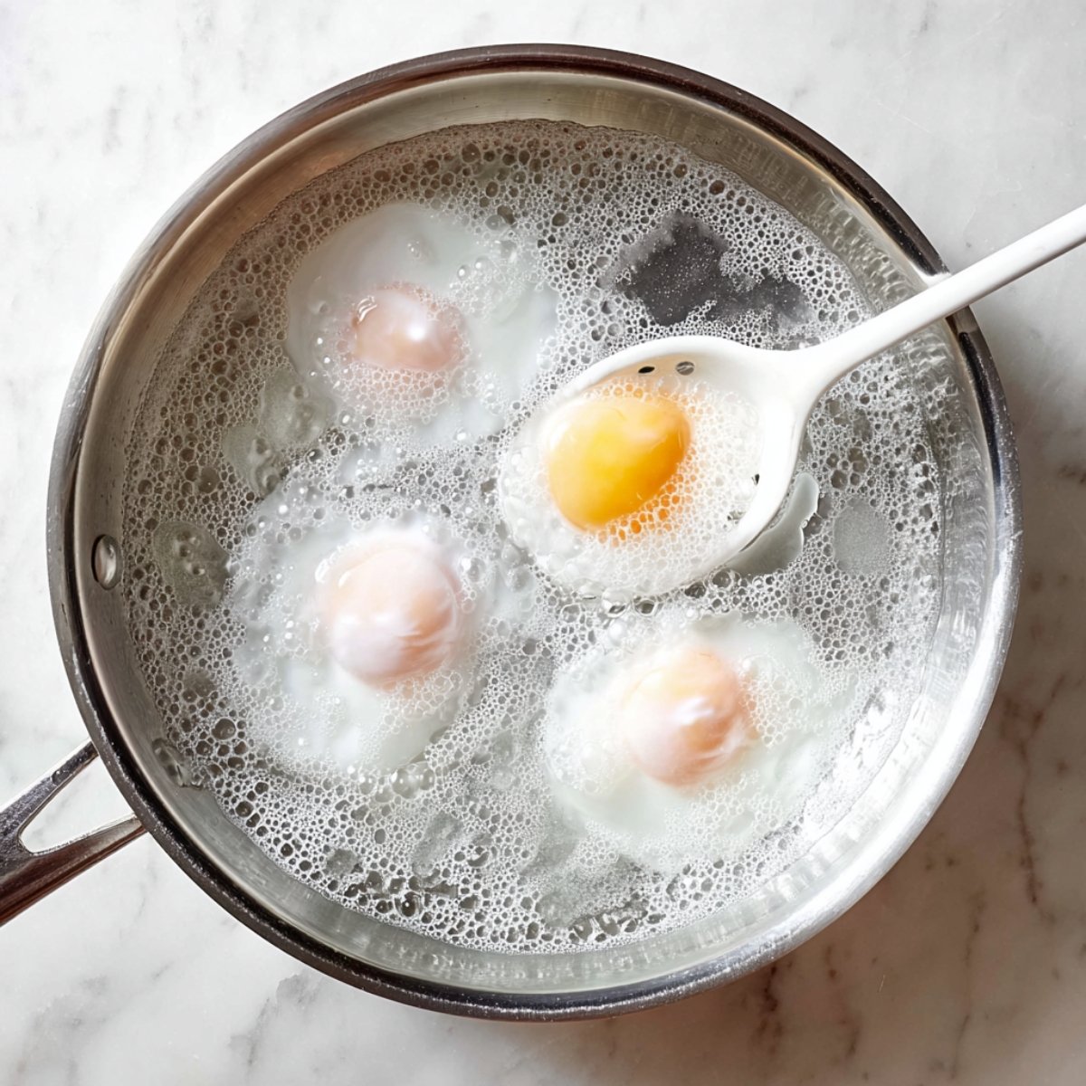 Four eggs poaching in simmering water in a metal pan, with a slotted spoon lifting one egg.