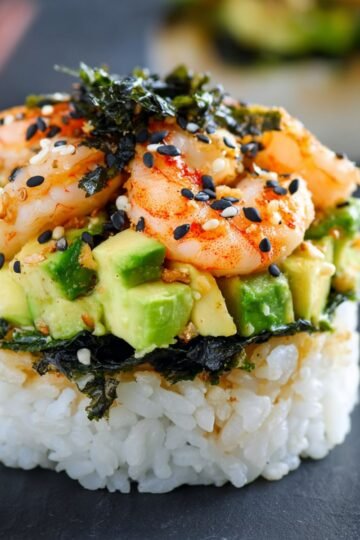 Close-up of a single portion of Salmon Sushi Bake on a dark surface, showing layers of rice, nori, diced avocado, and shrimp, garnished with black sesame seeds and chopped nori