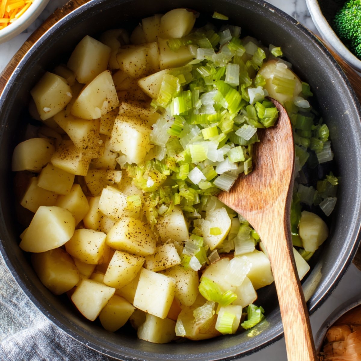 A close-up of a black pot containing cubed potatoes and chopped celery, lightly seasoned with black pepper, with a wooden spoon resting inside.