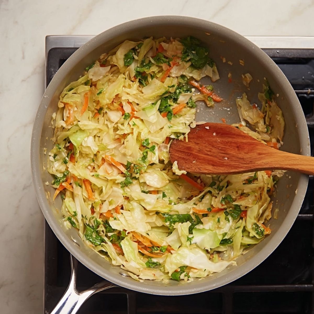 A top-down view of a frying pan on a stove containing sautéed cabbage mixed with shredded carrots and fresh herbs, being stirred with a wooden spatula.