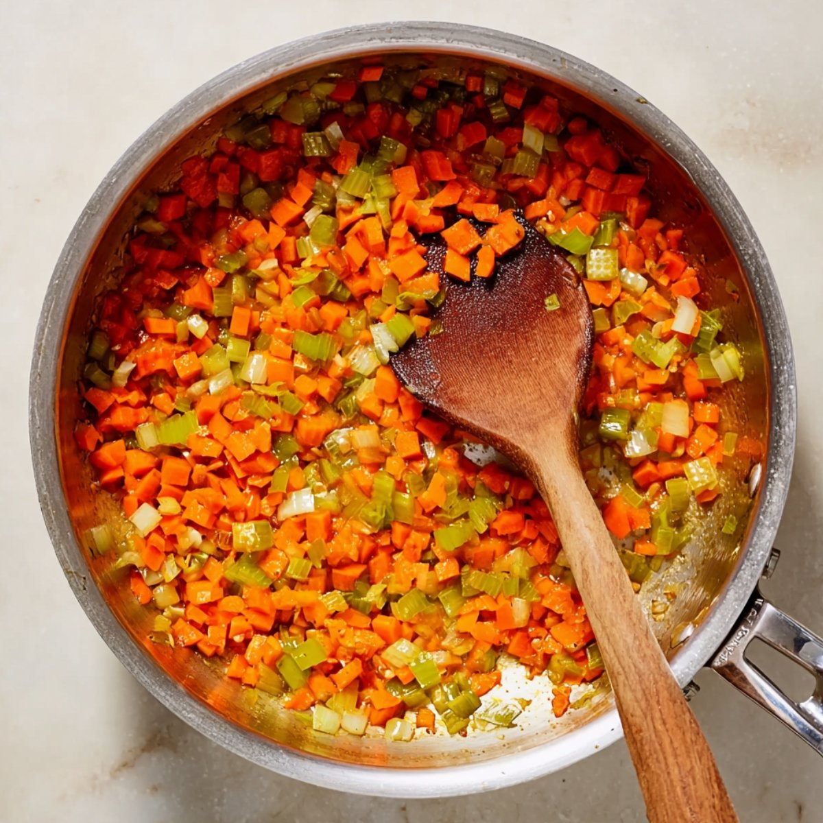 A saucepan filled with finely diced carrots and celery being sautéed, with a wooden spoon resting in the pan.