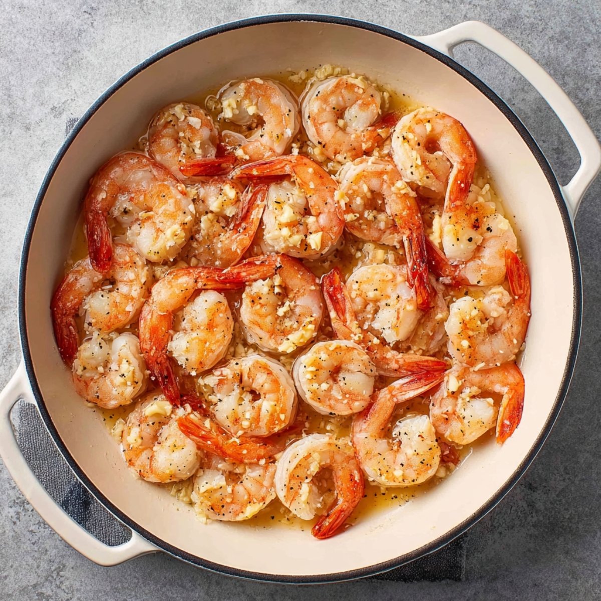 The same skillet now filled with pink, partially cooked shrimp arranged in a single layer, with garlic and butter mixture coating them.