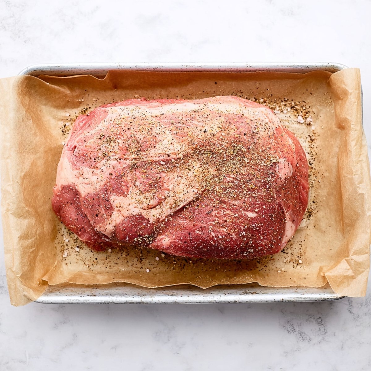 A raw pork roast on a parchment-lined baking sheet, seasoned generously with salt and black pepper.