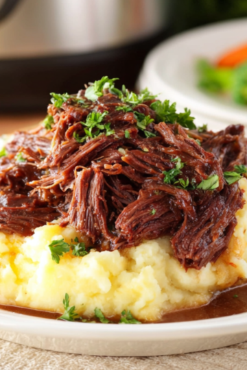 A plated serving of Slow Cooker Beef Manhattan in brown gravy over mashed potatoes, garnished with chopped parsley, with blurred vegetables in the background.