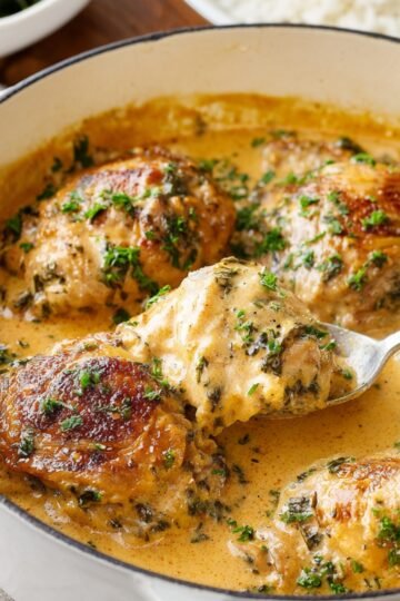 A skillet of smothered chicken in a creamy, herb-filled sauce, with one piece being lifted on a spoon, showing a golden-brown crust.