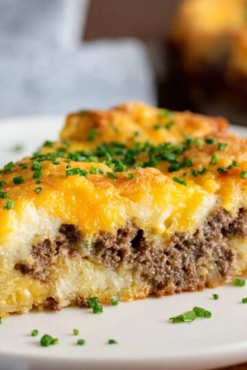 Close-up of a slice of baked tater tot breakfast casserole on a white plate, showing layers of ground beef, eggs, and melted cheddar cheese, garnished with chopped chives.