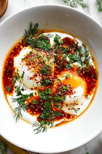 Bowl of Turkish eggs with poached eggs on yogurt, topped with chili oil, fresh herbs, and served with coarse salt on the side.
