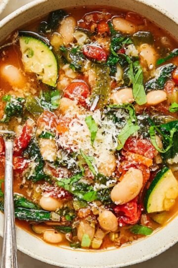A bowl of white bean and kale soup garnished with fresh herbs and Parmesan, with a spoon in the bowl, ready to serve.